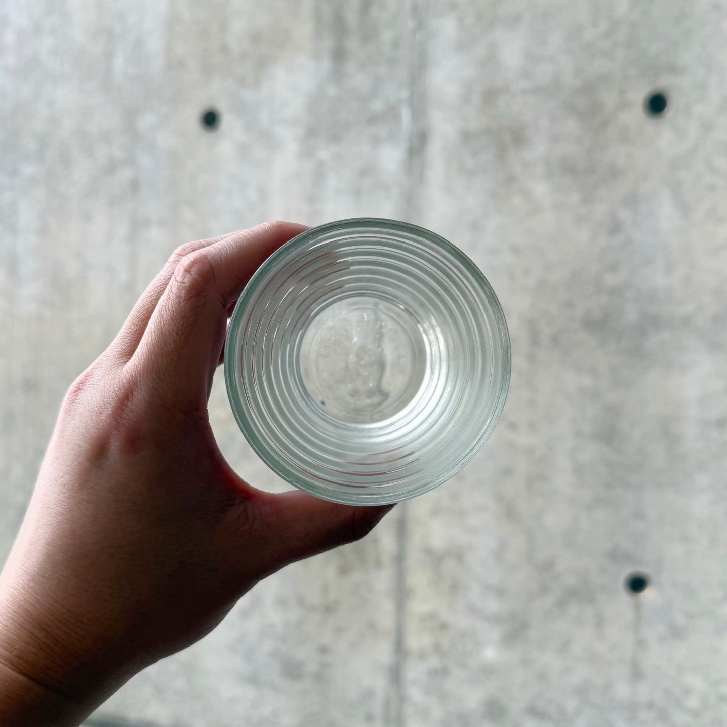 A hand holding the Aino Aalto Karhula-Iittala Bölgeblick Clear Glass Tumbler, showing its concentric circular pattern inside the clear glass against a neutral concrete background.