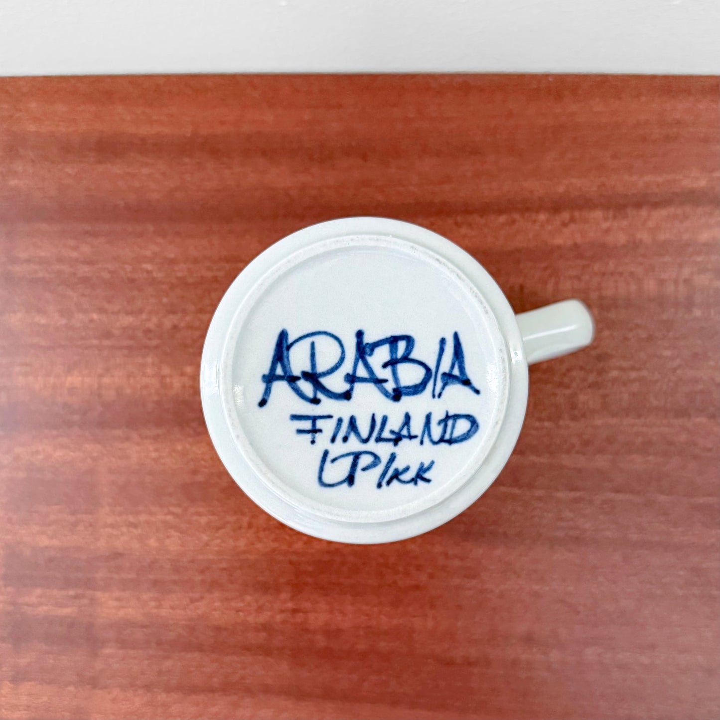 Bottom view of the Arabia Finland Anemone Cocoa Mug showing the blue handwritten maker's mark on white ceramic, on a wooden surface.