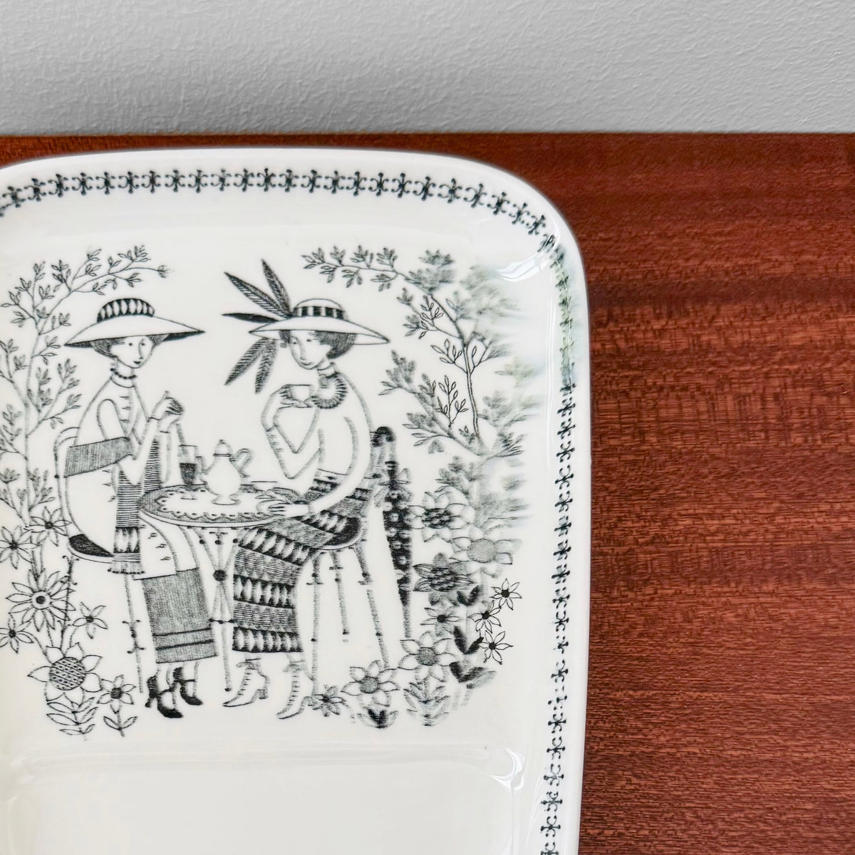 Close-up of a vintage ceramic plate with black and white illustration of two women in hats sitting at a table, part of the Arabia Finland Emilia TV Set displayed on a wooden surface.