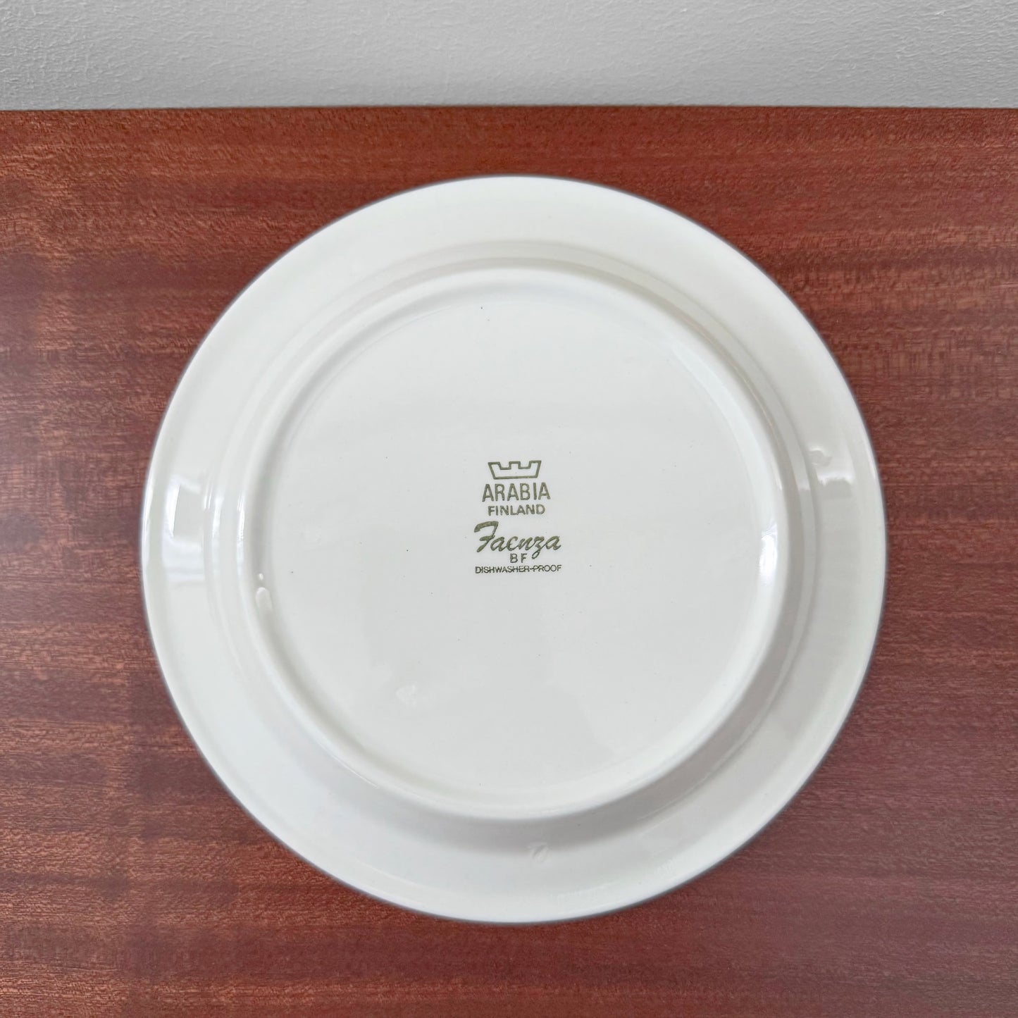 Back view of a white ceramic cake plate from the Arabia Finland Faenza Brown Tea Cup Trio Set showing the maker's mark and vintage design logo on a wooden surface.