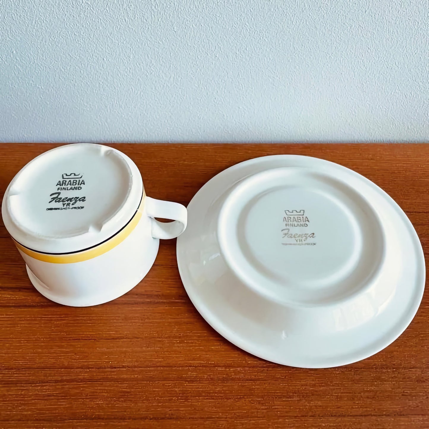 Arabia Finland Faenza Yellow Tea Cup with Saucer showing the bottom marks on white ceramic with a yellow stripe, displayed on a wooden surface in excellent vintage condition.