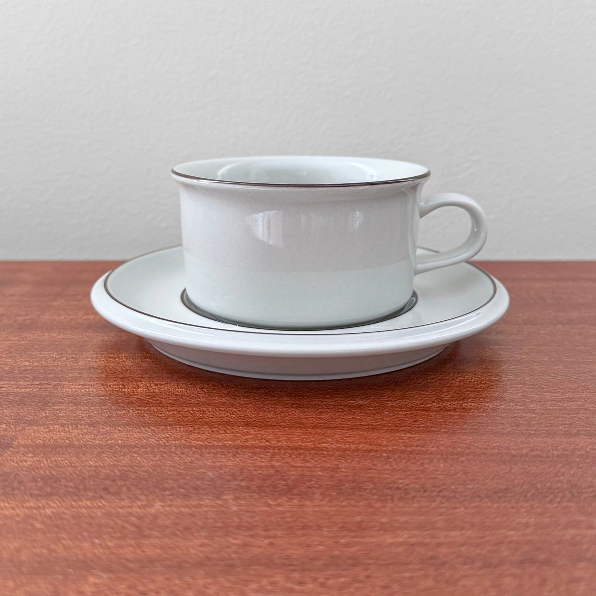 Arabia Finland Fennica Tea Cup with Saucer in white ceramic featuring a minimalist design with fine silver trim, displayed on a wooden surface against a plain background.