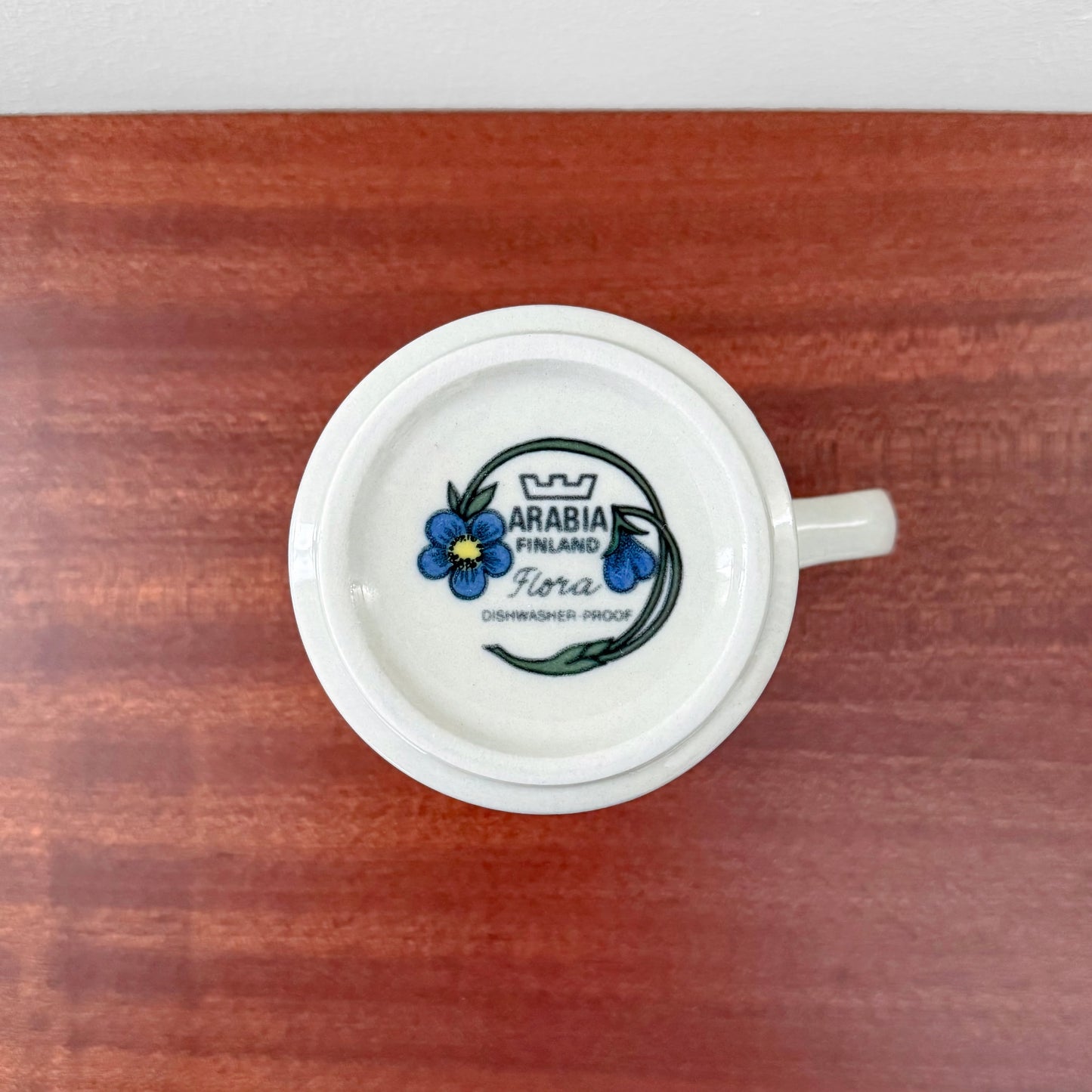 Bottom view of an Arabia Finland Flora Cocoa Mug showing the vintage mark with blue floral design on white ceramic, placed on a wooden surface.