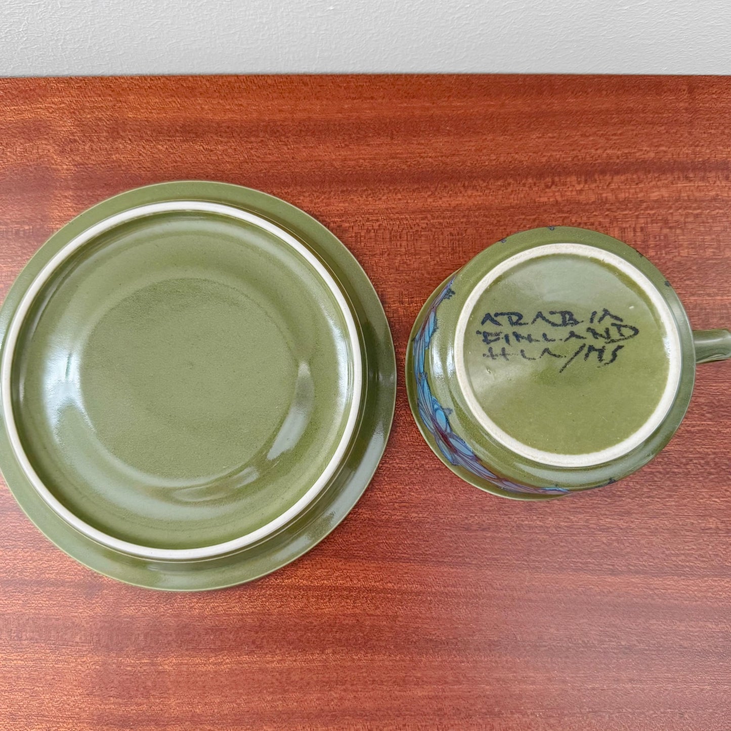 Arabia Finland Hilkka-Liisa Ahola Green Tea Cup with Saucer showing the green ceramic cup with hand-painted signature on the underside and matching saucer on a wooden surface.
