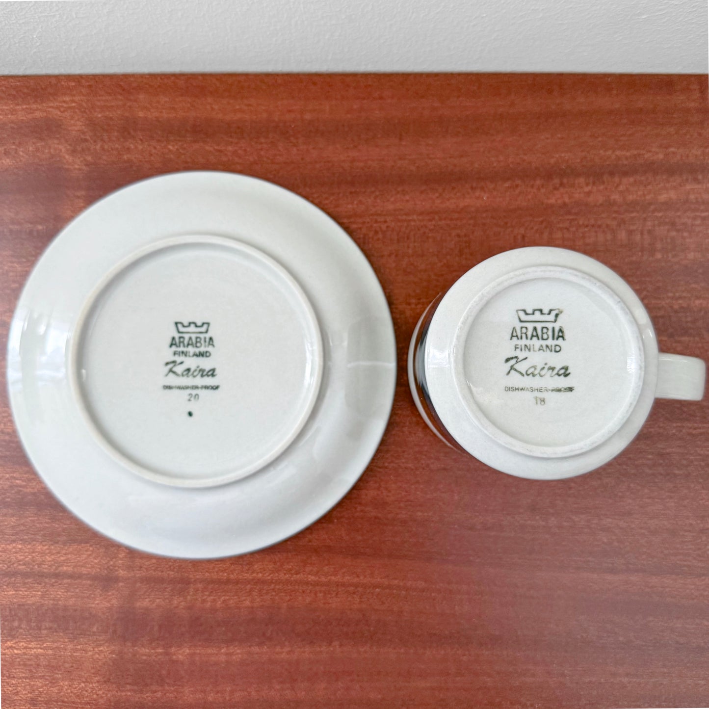 Bottom view of the Arabia Finland Kaira Tea Cup with Saucer showing the vintage maker’s marks on white ceramic pieces placed on a wooden surface.