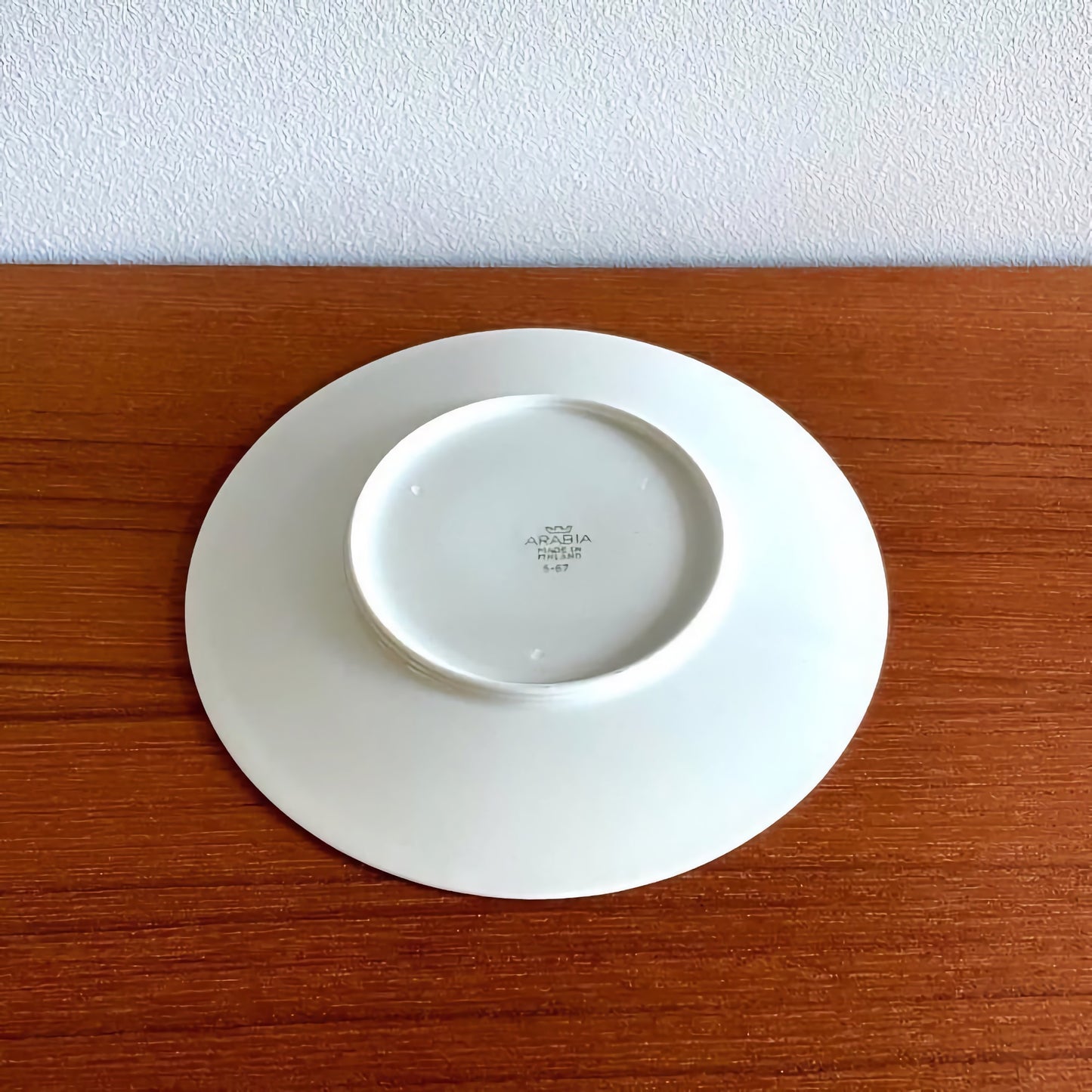 Back view of a white ceramic plate from the Arabia Finland Katinka Espresso Cup Trio Set, showing the maker's mark on a wooden surface.