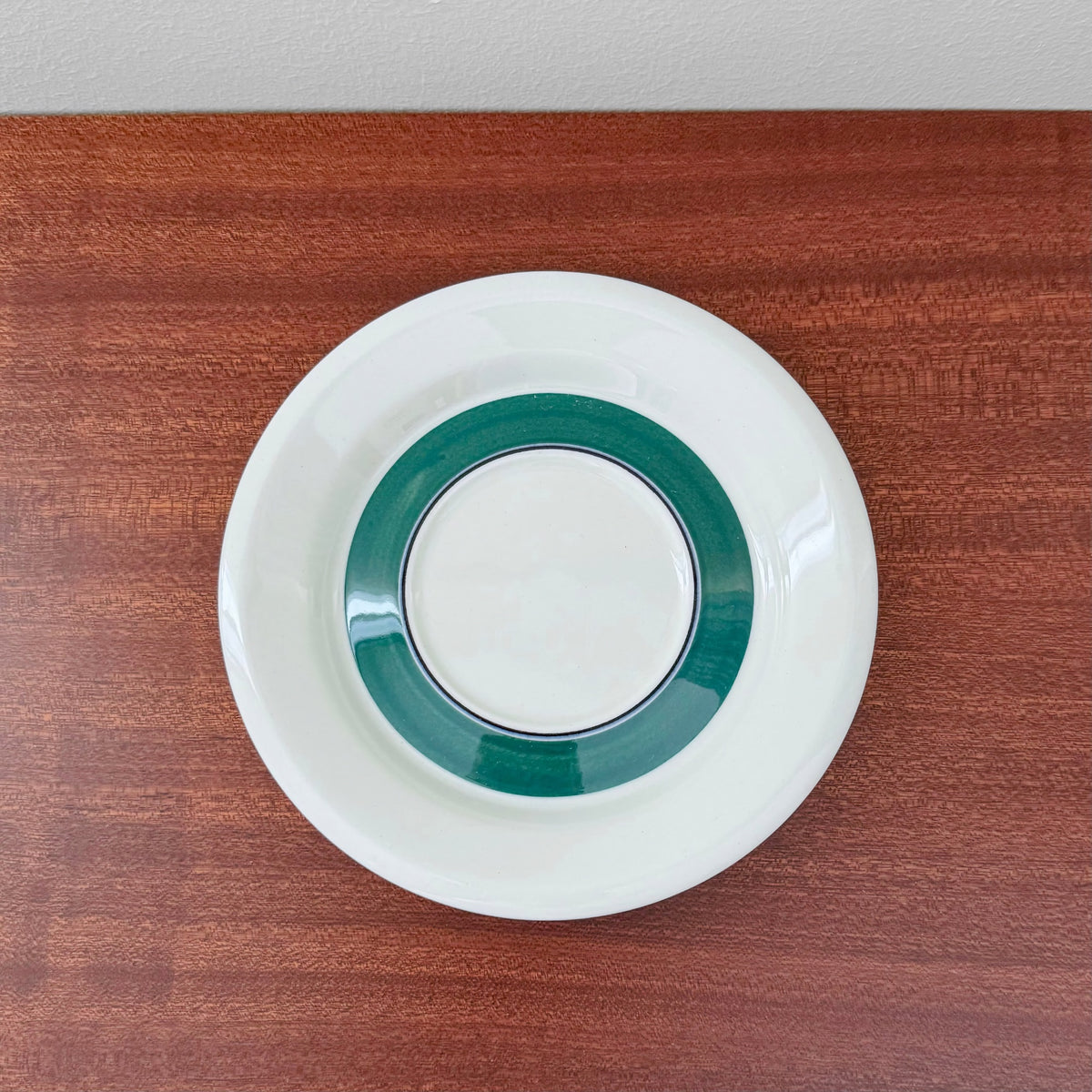 A white ceramic saucer with a wide green band near the center, part of the Arabia Finland Kirsikka Coffee Cup Trio Set, displayed on a wooden surface.