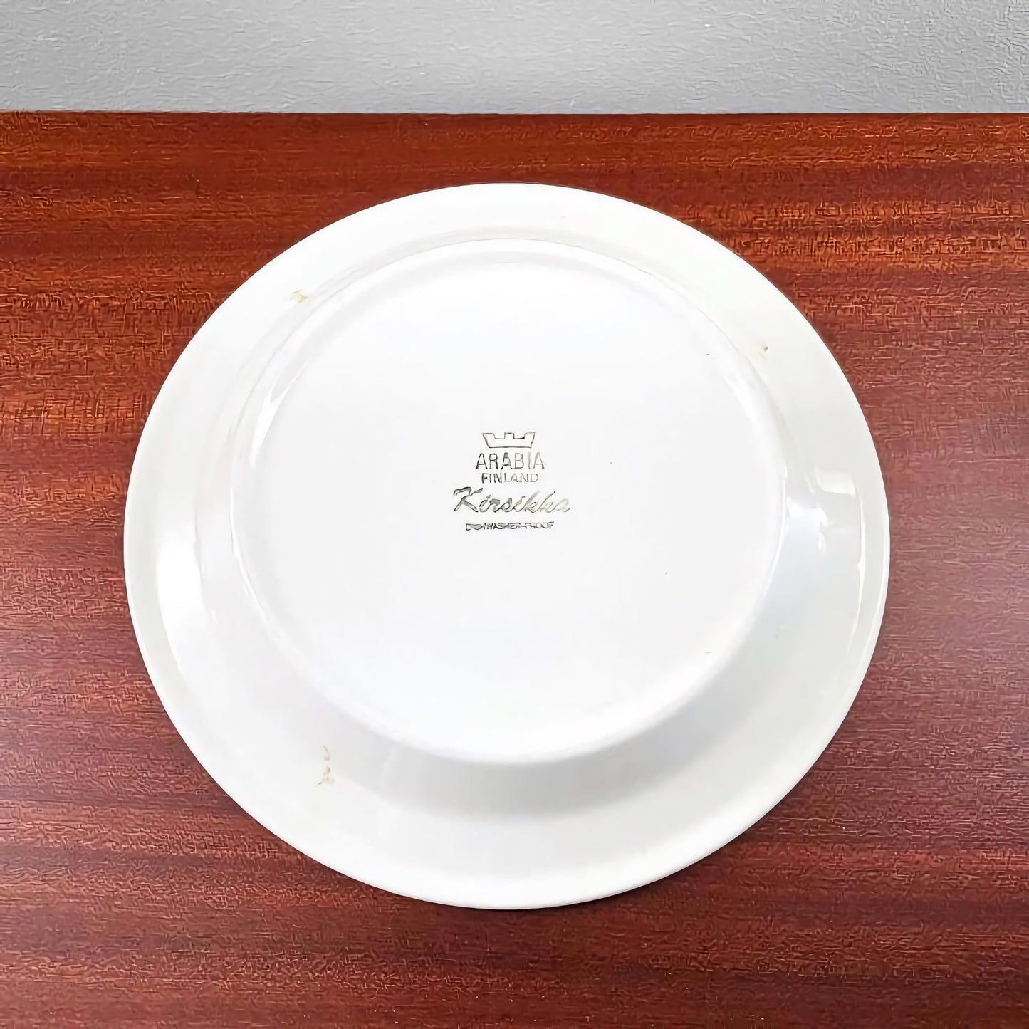Backside view of a white ceramic plate from the Arabia Finland Kirsikka Tea Cup Trio Set, showing the vintage maker's mark on the base against a wooden surface.