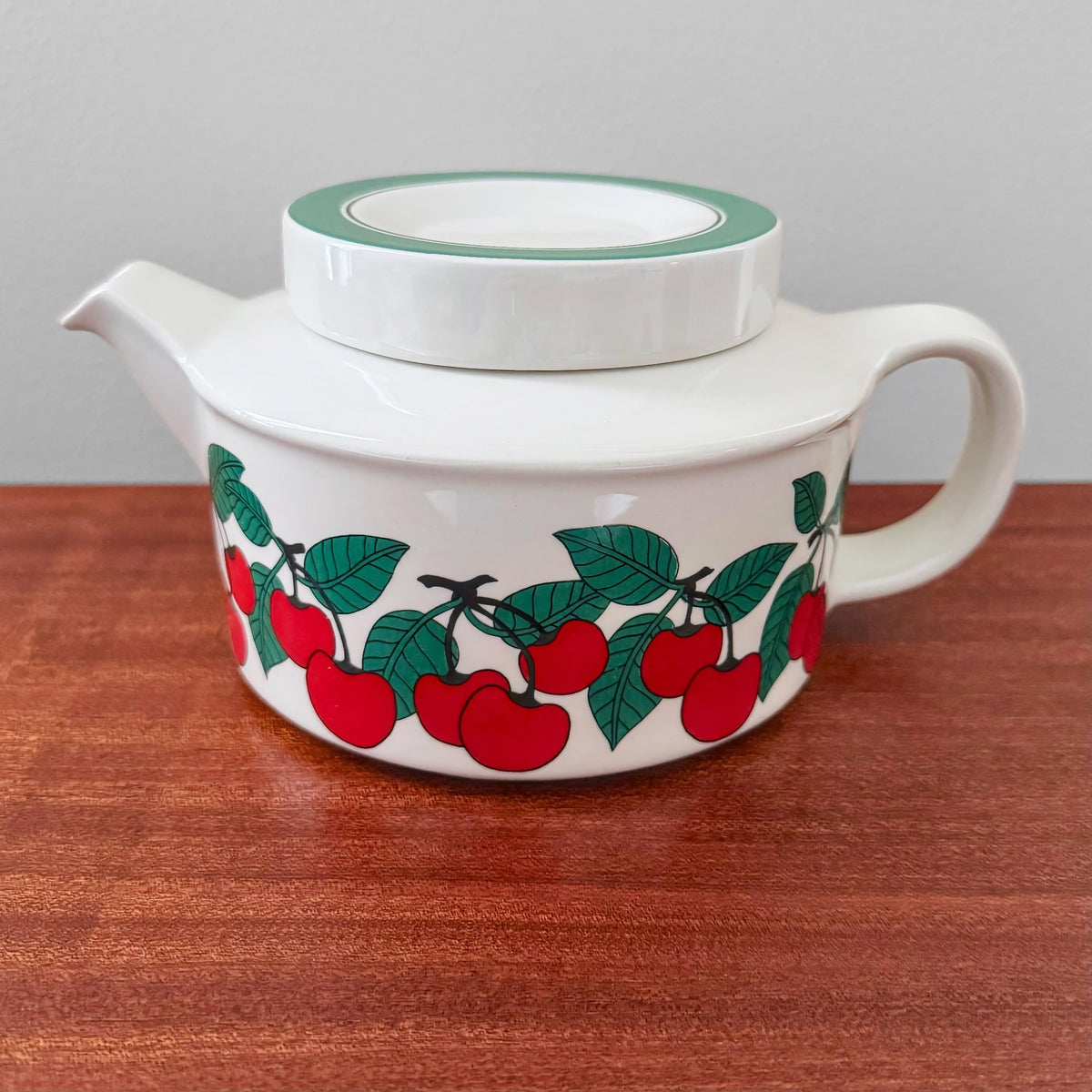 Arabia Finland Kirsikka Teapot featuring a white ceramic body with a green rim lid and vibrant red cherry and green leaf pattern around the base, vintage Nordic design.