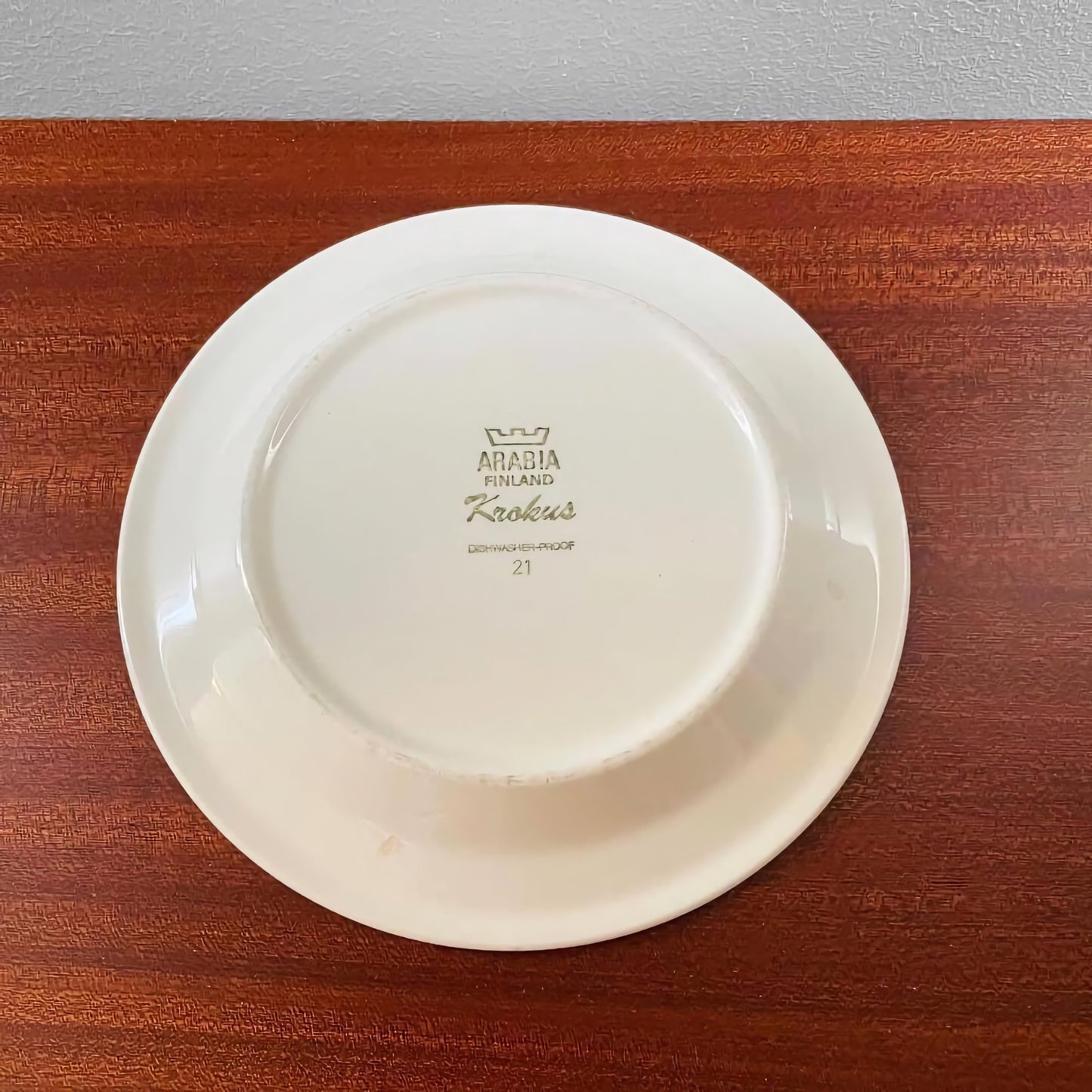 Bottom view of a white ceramic plate marked with Arabia Finland Krokus branding, placed on a wooden surface, part of the Arabia Finland Krokus Coloured Espresso Cup Trio Set.