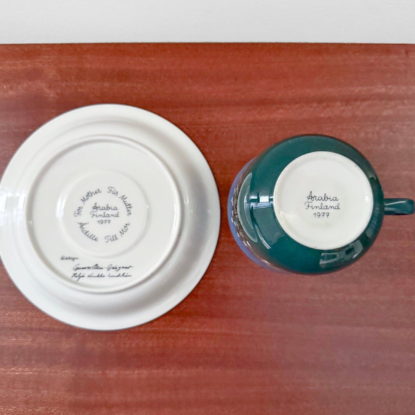 Bottom view of Arabia Finland Mother's Cup 1977 showing the white ceramic base of the saucer and the green ceramic base of the cup with maker's marks on a wooden surface.