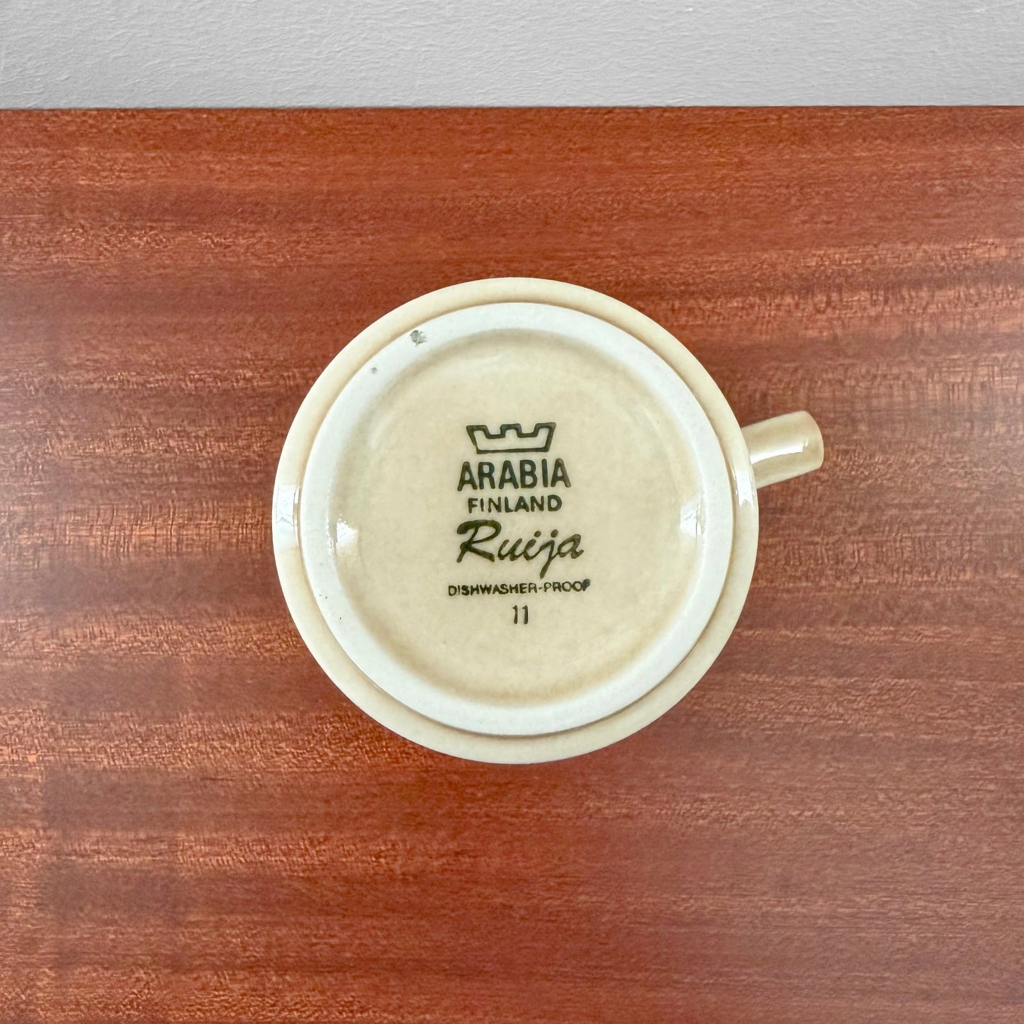 Bottom view of a vintage ceramic Arabia Finland Ruija Cocoa Mug showing maker’s mark and dishwasher-proof label on a wooden surface.