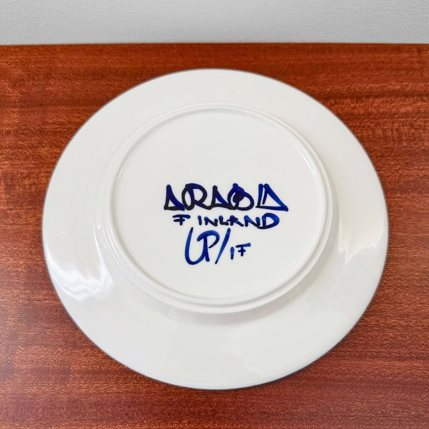 Bottom view of a white ceramic plate from the Arabia Finland Valencia Tea Cup Trio Set with the hand-painted Arabia Finland maker's mark and signature on the underside, placed on a wooden surface.