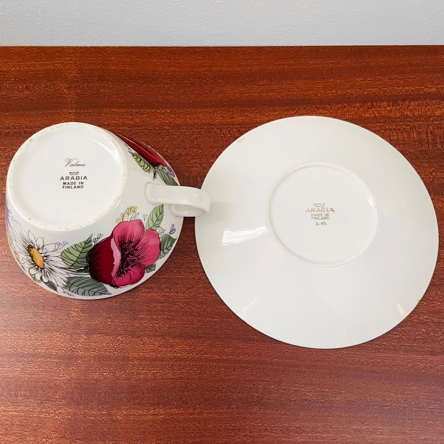 Arabia Finland Valmu Tea Cup with Saucer showing the floral design on the cup’s exterior and the maker’s marks on the bottom of both pieces, displayed on a wooden surface.