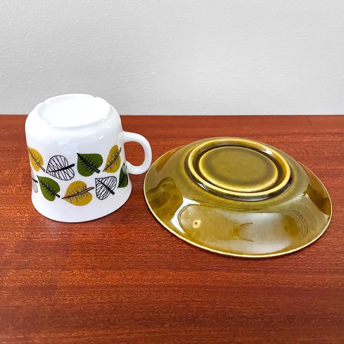 Upsala-Ekeby/Gefle Bladranka Coffee Cup with Saucer shown upside down, featuring a white ceramic cup with green and yellow leaf patterns and a glossy olive green saucer.