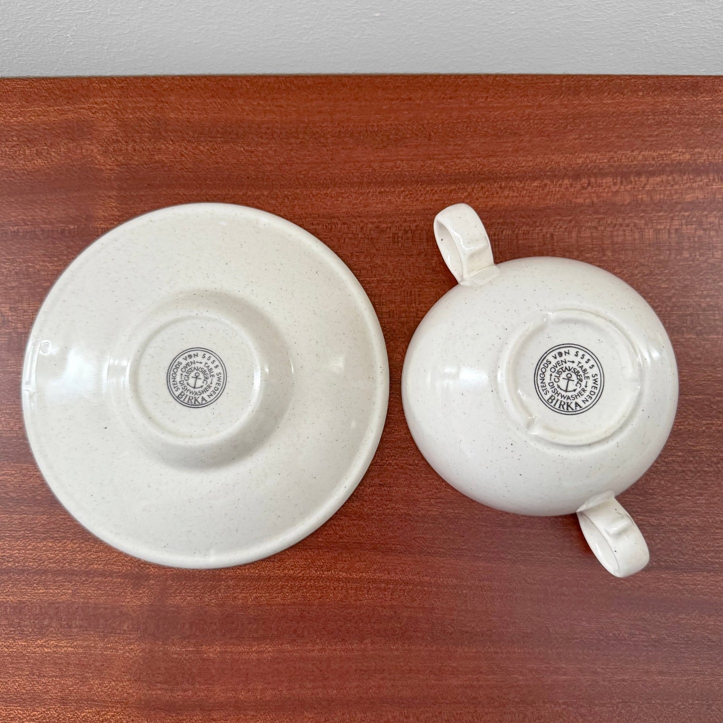 Gustavsberg Birka Bouillon Cup with Saucer shown upside down on a wooden surface, featuring a creamy off-white ceramic finish and vintage maker’s mark designed by Stig Lindberg.
