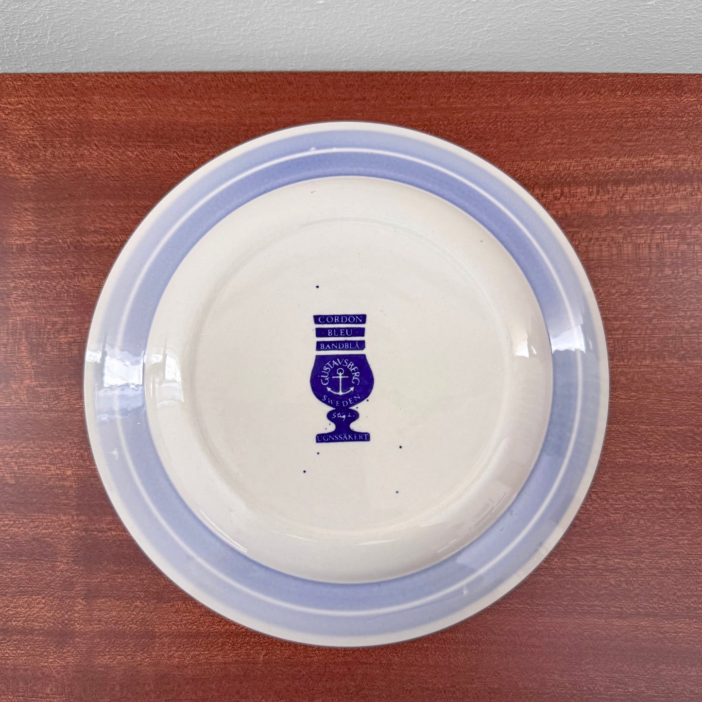 Back of a cake plate with light blue border and Gustavsberg Cordon Bleu Tea Cup Trio Set mark in dark blue on white ceramic surface.
