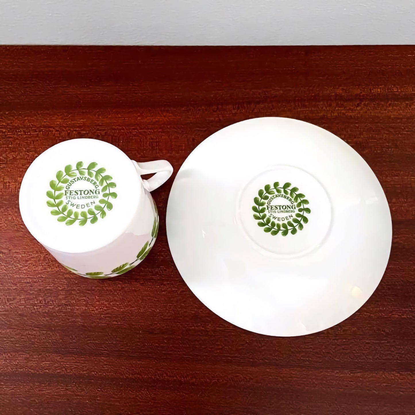 Gustavsberg Festong Coffee Cup with Saucer shown upside down on a wooden surface featuring vintage Scandinavian ceramic design with a green laurel wreath pattern and maker’s mark.