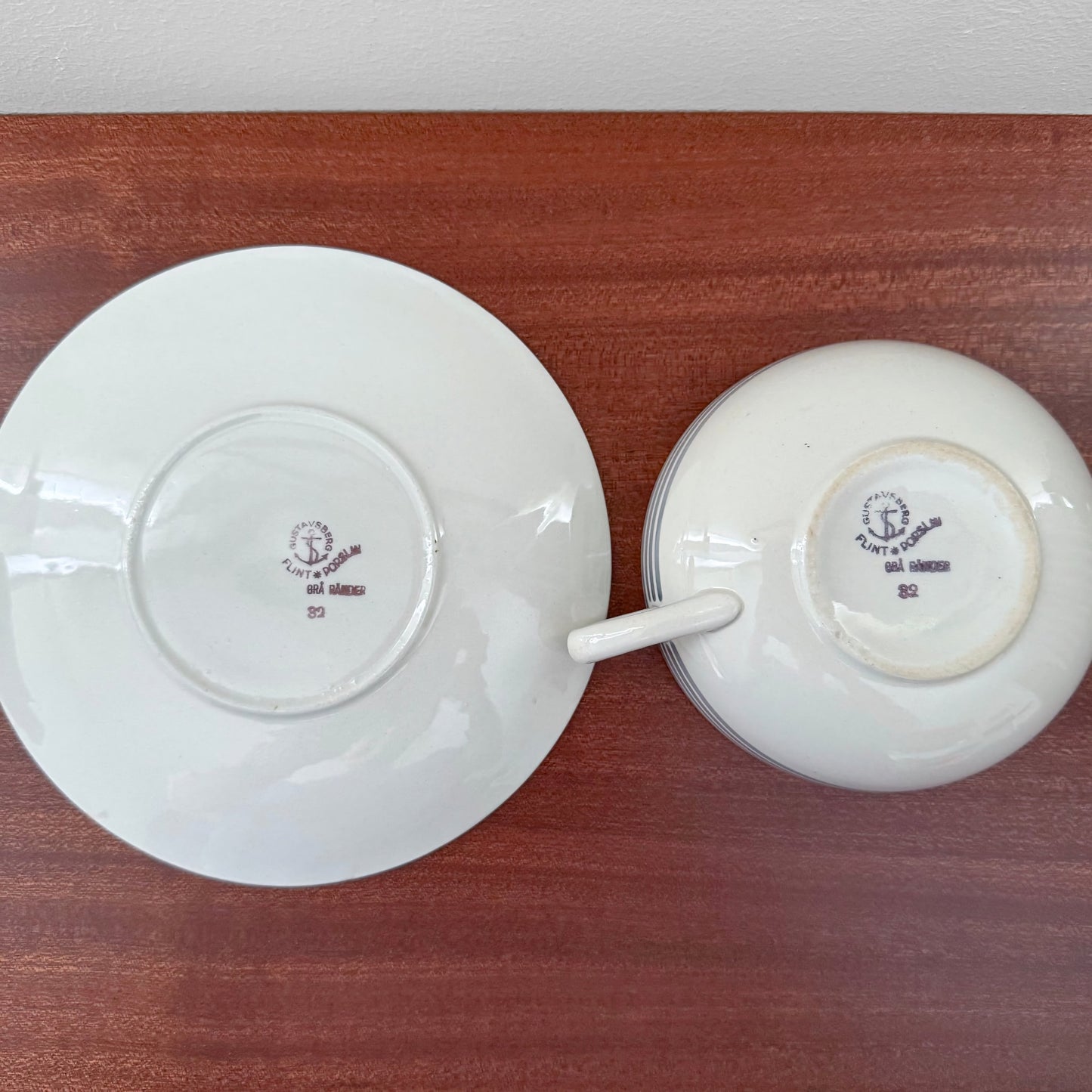 Gustavsberg Grå Ränder Tea Cup with Saucer shown from the bottom, displaying vintage maker’s marks on the ceramic in white and purple, placed on a wooden surface.