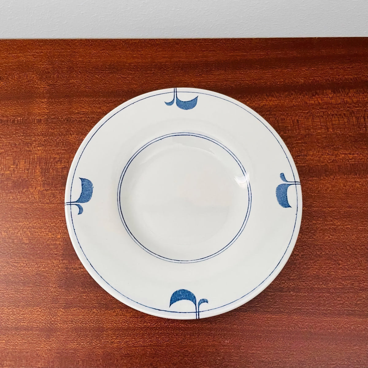 Gustavsberg Ming Tea Cup with Saucer showing a white ceramic saucer with simple blue floral motifs and double line detailing on a wooden surface, designed by Stig Lindberg.