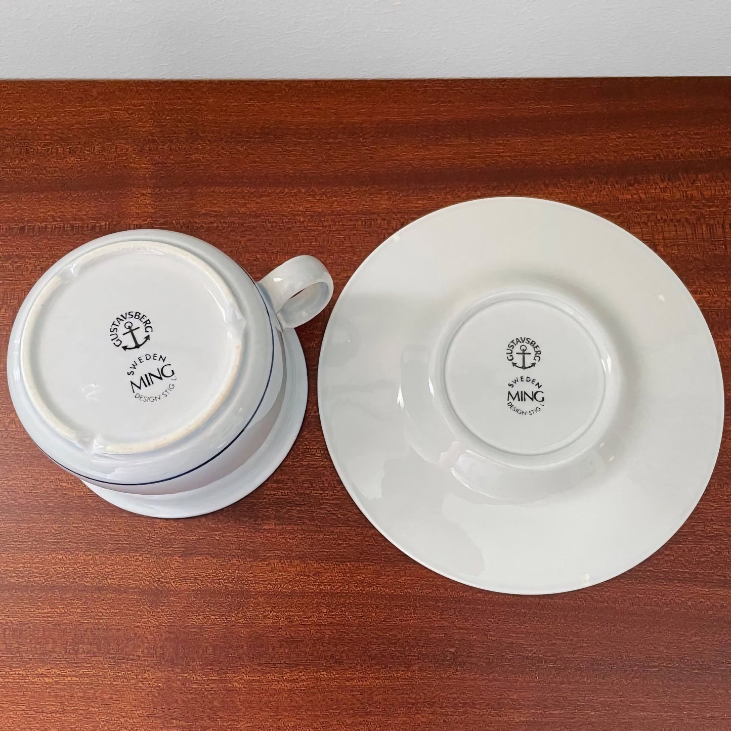 Gustavsberg Ming Tea Cup with Saucer in white ceramic, showing the underside markings with Gustavsberg Sweden and Ming logos on a wooden surface.