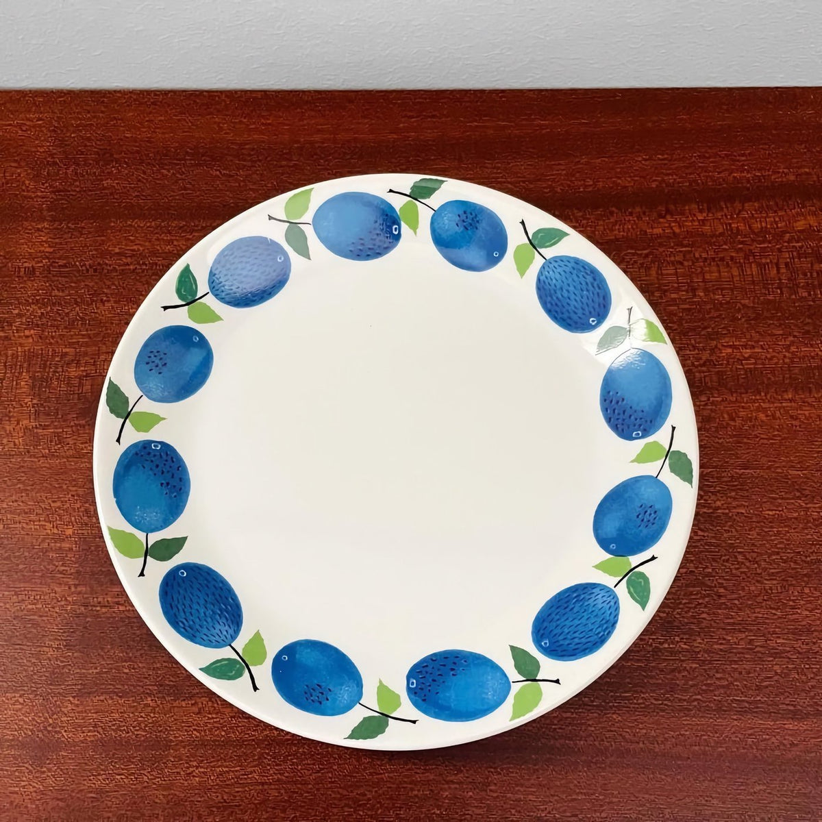 Gustavsberg Prunus Cake Plate featuring a circular pattern of blue plums with green leaves on a white ceramic base, showcasing vintage Scandinavian design by Stig Lindberg.