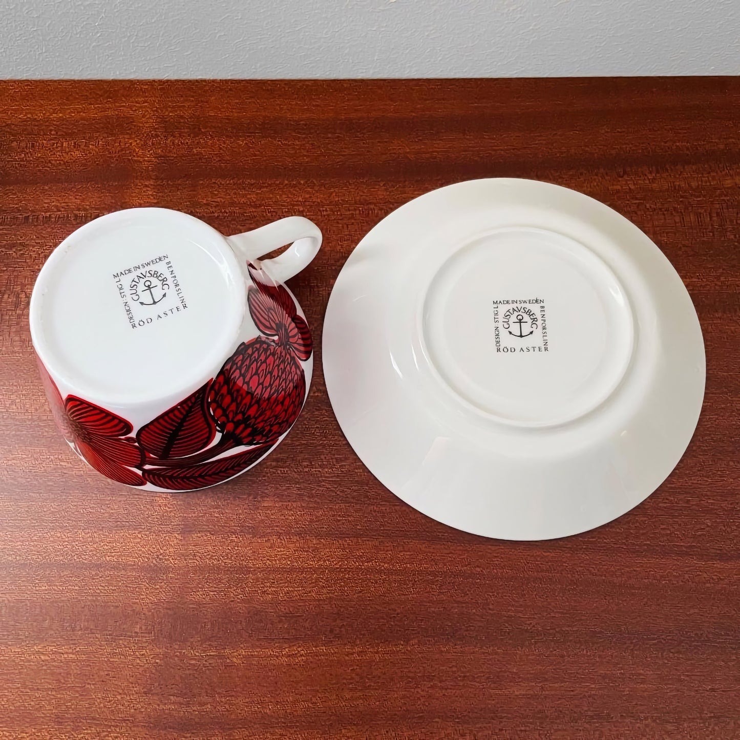 Gustavsberg Röd Aster Tea Cup with Saucer showing the underside stamps, featuring red floral Nordic ceramics designed by Stig Lindberg placed on a wooden surface.