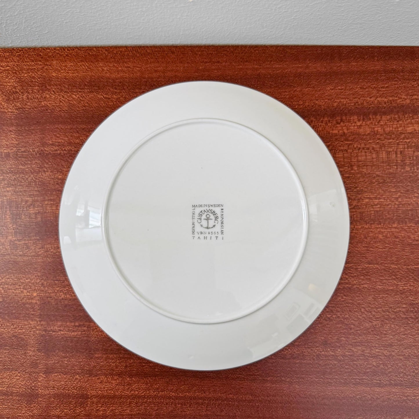 Back view of a white cake plate from the Gustavsberg Tahiti Coffee Cup Trio Set, featuring the stamped maker's mark by Stig Lindberg, placed on a wooden surface.