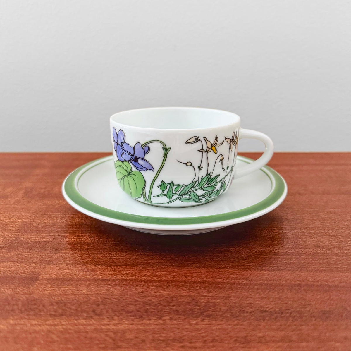 IKEA Midsommar Tea Cup with Saucer featuring a vintage floral design in blue, green, and yellow on white ceramic, displayed on a matching white saucer with green rim.
