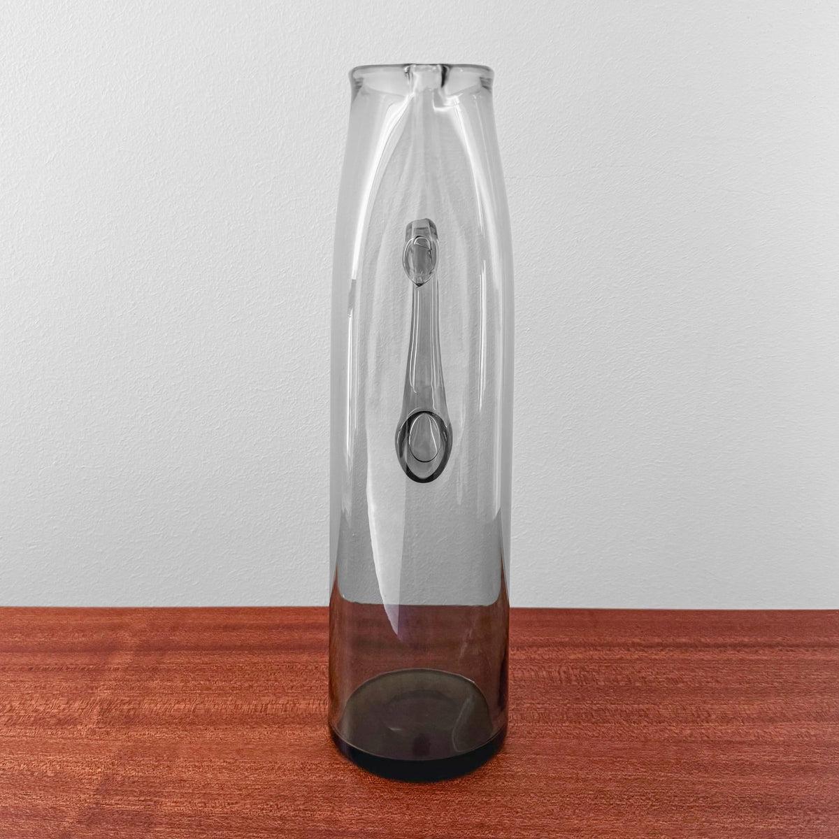 Ingeborg Lundin Orrefors Grey Glass Pitcher standing on a wooden surface against a white wall, showcasing its tall, slender form and translucent smoky grey glass design.