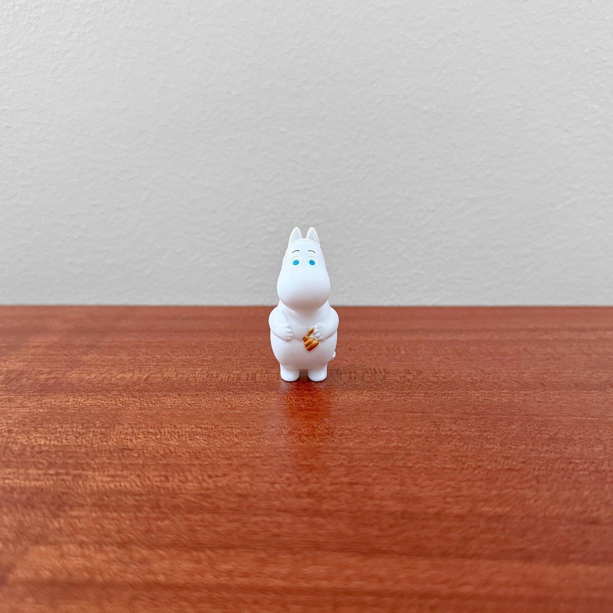 Small white figurine character with blue eyes holding a small item, displayed on wooden surface, part of Moomin Miniature Lamp Part 3 Capsule Toys collection.