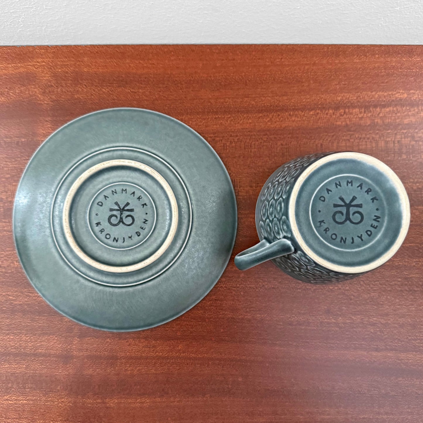 Quistgaard Azur Tea Cup with Saucer showing the underside with embossed Nordic Kronjyden Denmark marks on textured ceramic surfaces in a deep blue-green glaze on a wooden table.