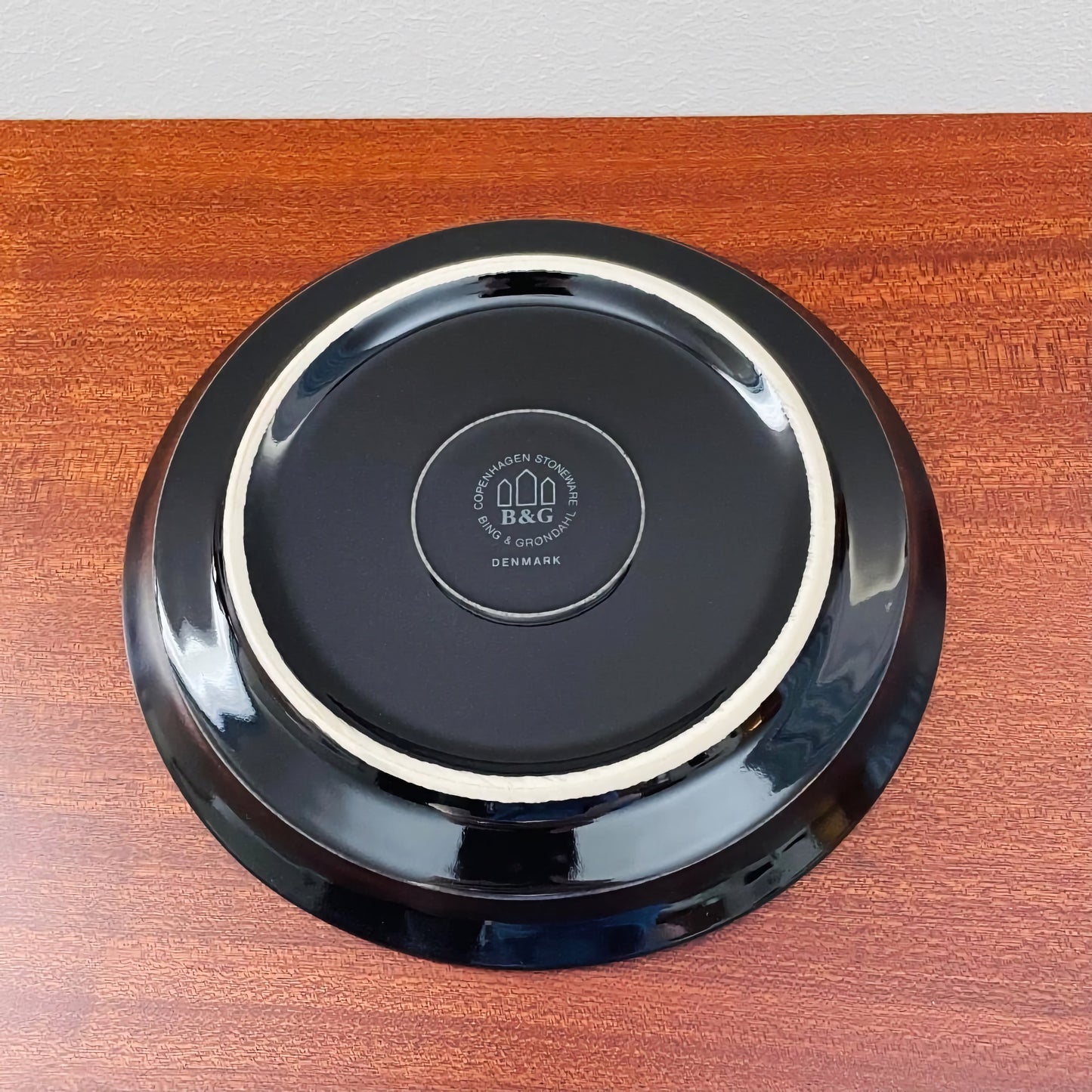 Bottom view of a black ceramic saucer from the Quistgaard Cordial Palet Black Tea Cup Trio Set, showing the Bing & Grøndahl maker's mark on a polished wooden surface.