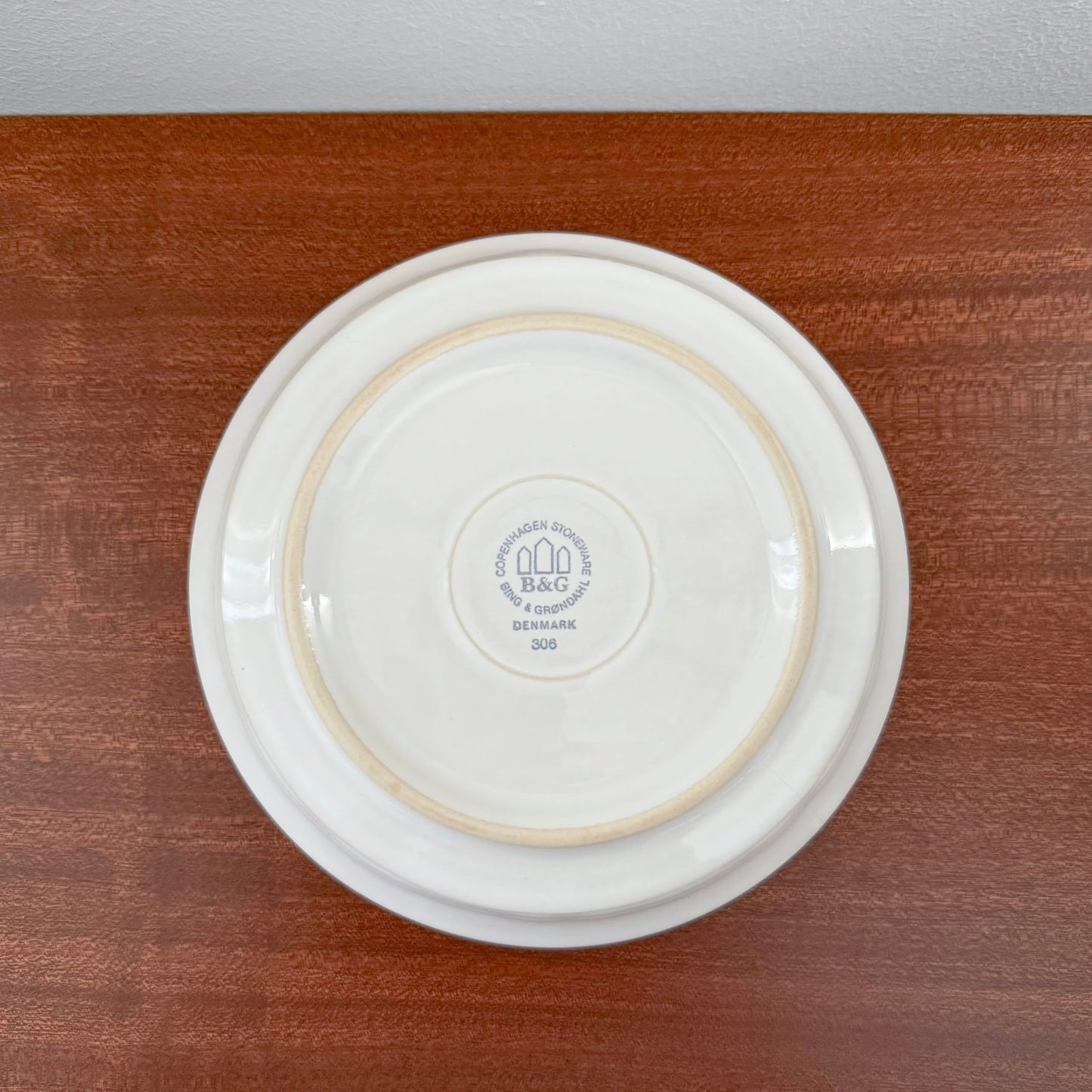 Bottom view of a white ceramic plate from the Quistgaard Cordial Palet White Tea Cup Trio Set, showing the Bing & Grøndahl maker’s mark on a wooden surface.
