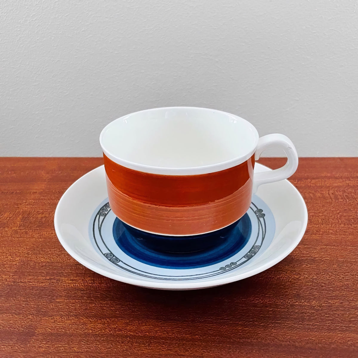 Rörstrand Augusta Tea Cup with Saucer featuring a white cup with a bold reddish-brown band and a white saucer decorated with blue and gray circular patterns, in vintage Nordic ceramic style.
