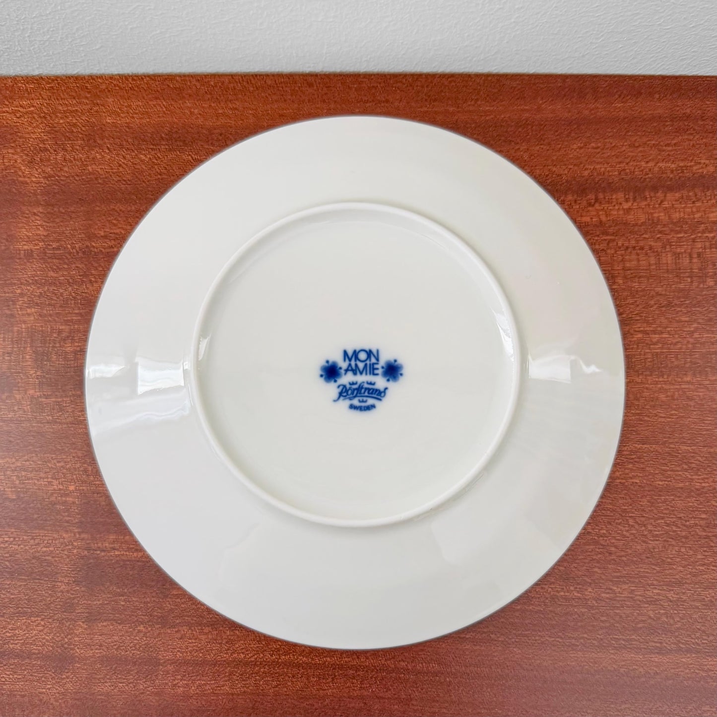 Back view of a white ceramic plate from the Rörstrand Mon Amie Tea Cup Trio Set showing the blue Mon Amie and Rörstrand Sweden maker's marks on a wooden surface.