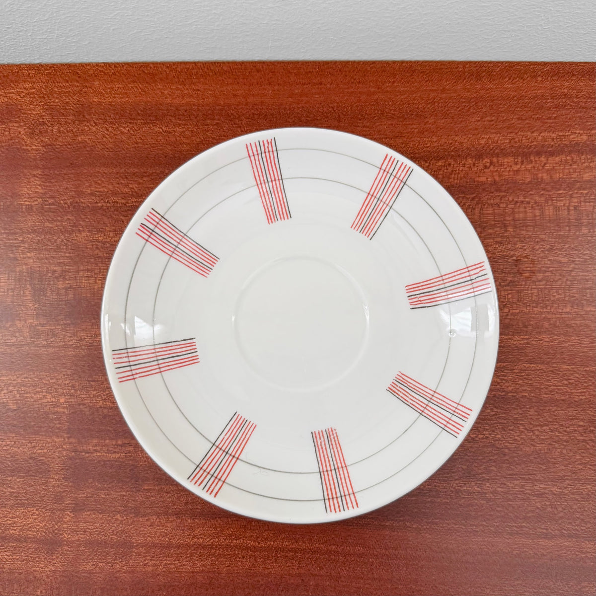 White ceramic saucer from the Rörstrand Striped Tea Cup Trio Set featuring red and black stripe patterns, placed on a wooden surface.