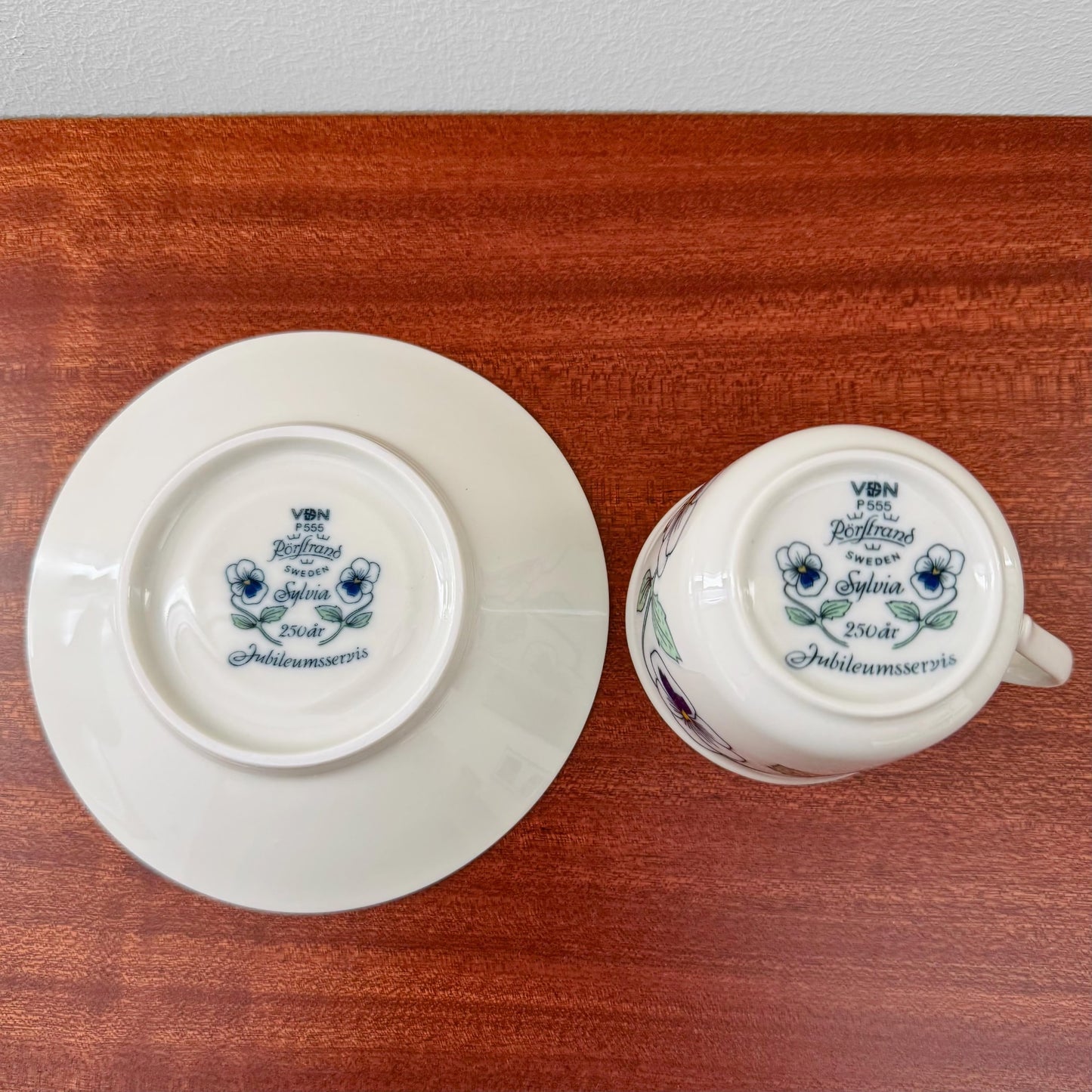 Rörstrand Sylvia Coffee Cup with Saucer showing the underside with blue floral design and commemorative text on white ceramic, placed on a wooden surface.