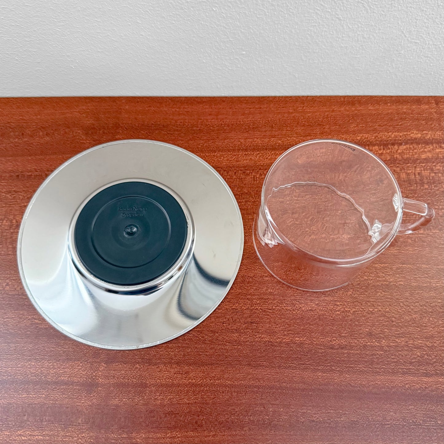Signe Persson-Melin Boda Nova Glass Coffee Cup with Steel Saucer displayed on a wooden surface, featuring a clear glass cup and polished steel saucer with a black base.