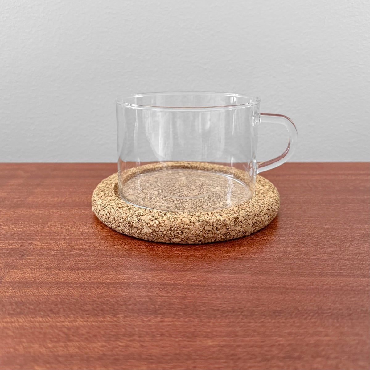 Signe Persson-Melin Boda Nova Glass Tea Cup with Cork Coaster featuring a clear glass cup with handle resting on a round natural cork coaster, displayed on a wooden surface.