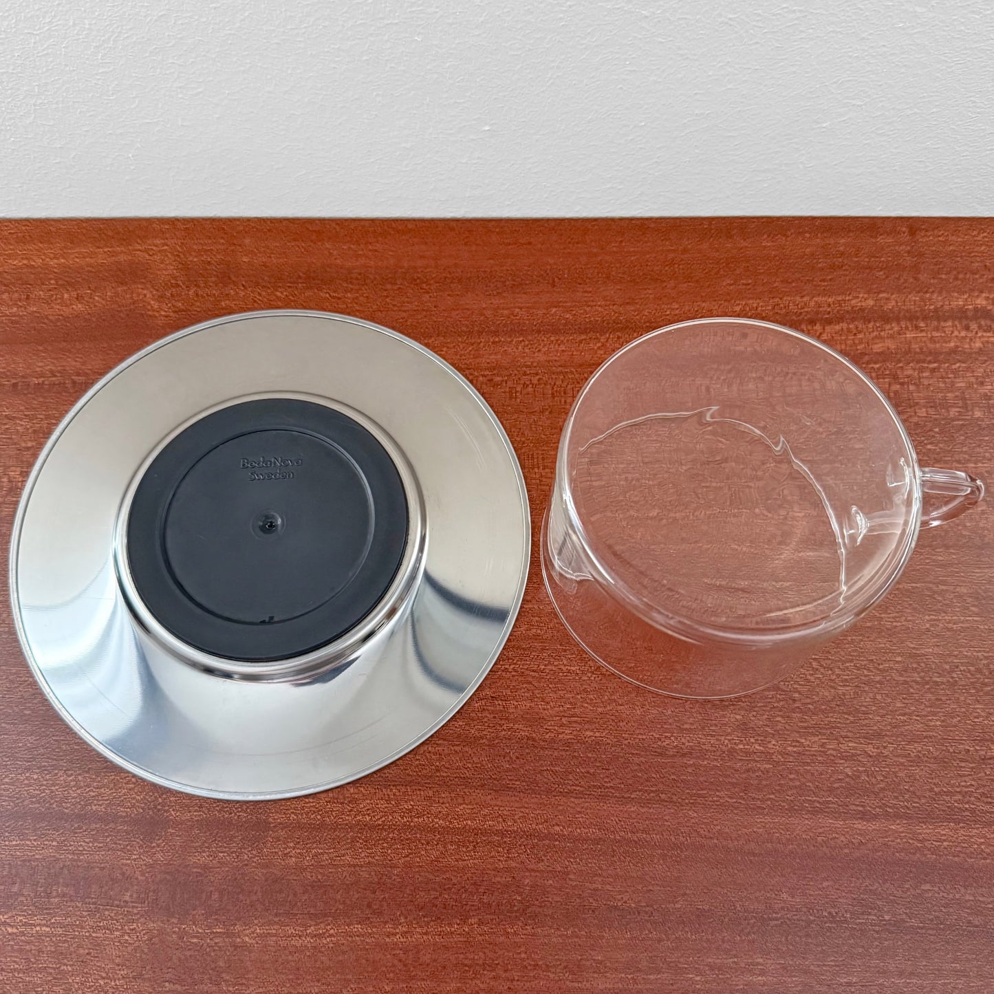 Signe Persson-Melin Boda Nova Glass Tea Cup with Steel Saucer showing a clear glass cup and a reflective stainless steel saucer placed on a wooden surface.