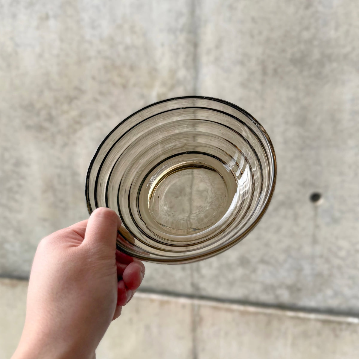 A hand holding the Aino Aalto Karhula Bölgeblick Smoke Glass Bowl (14.5 cm), a vintage Scandinavian decorative glass bowl with concentric rings and a subtle smoky tint.