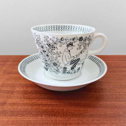 Arabia Finland Emilia Large Cocoa Cup with Saucer featuring a vintage black and white illustration of a seated figure among floral patterns, set on a wooden surface.