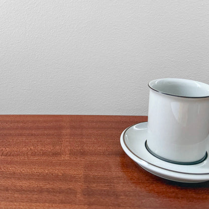 Arabia Finland Fennica Coffee Cup with Saucer in white ceramic with a simple dark rim design, placed on a wooden surface against a light gray wall.