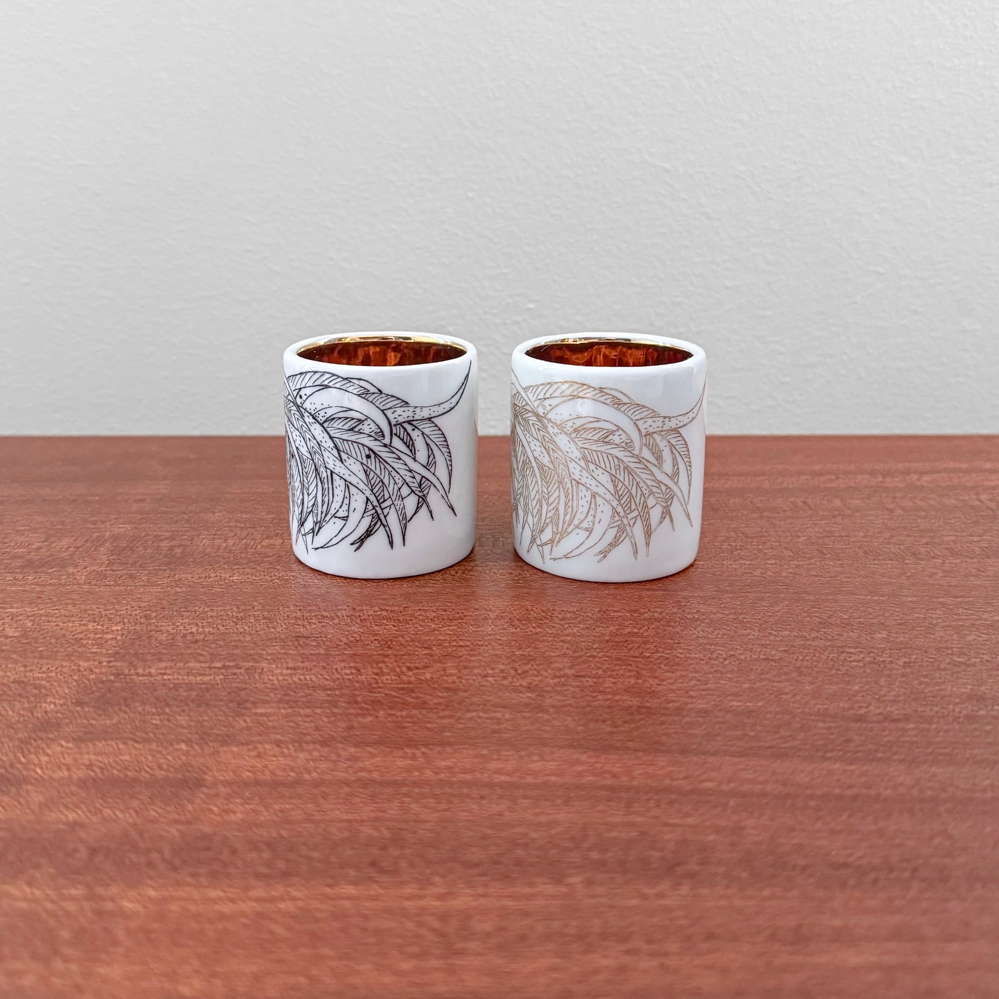 Pair of Arabia Finland Kukko Egg Cups featuring vintage hand-painted botanical designs in black and gold on white ceramic with a glossy finish, displayed on a wooden surface.