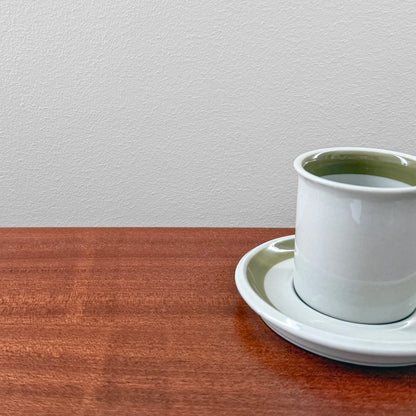 Arabia Finland Oliivi Coffee Cup with Saucer in white ceramic with olive green interior detail, displayed on a wooden surface against a plain light wall.