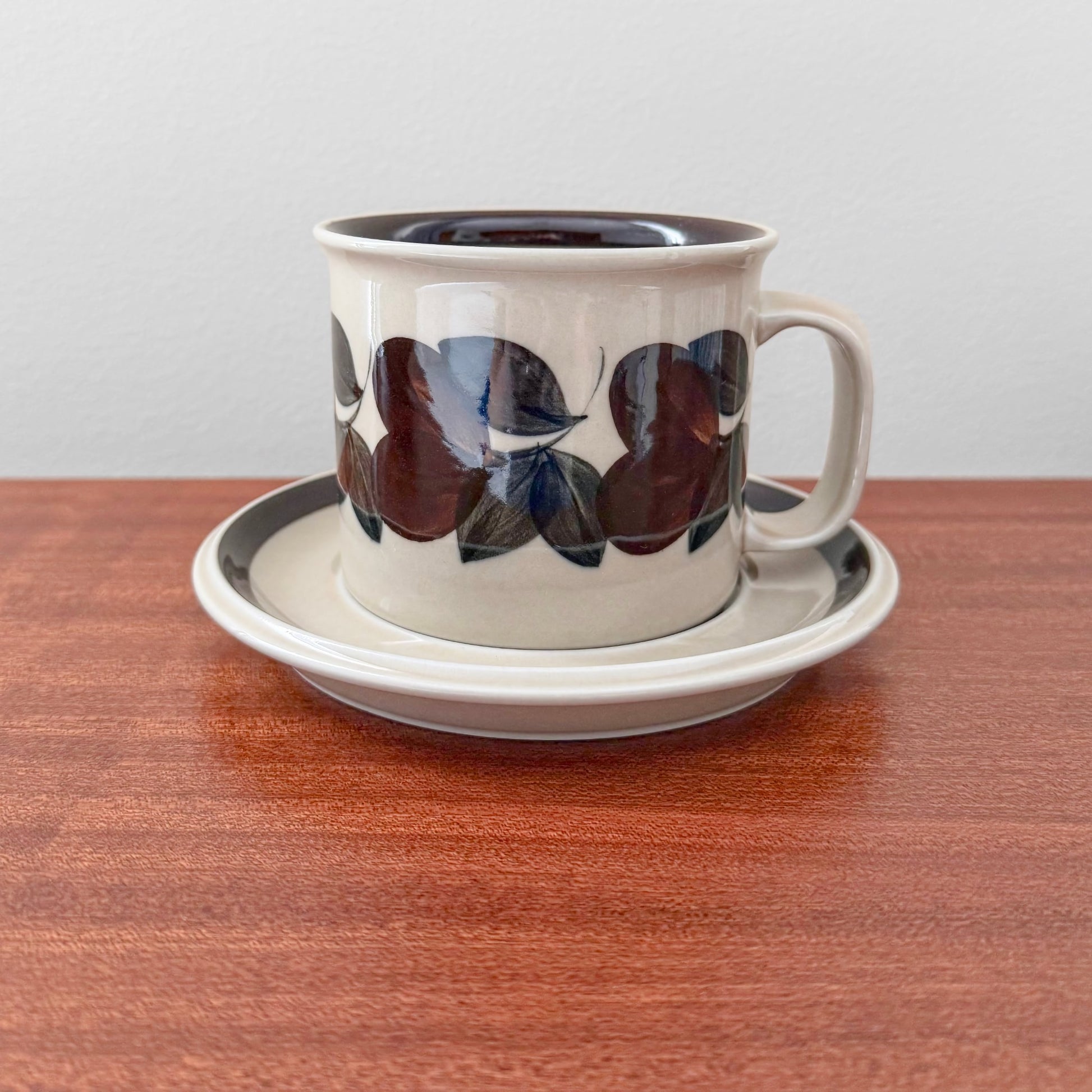 Arabia Finland Ruija Large Mug with Saucer featuring vintage ceramic design with dark leaf patterns on cream background, displayed on wooden surface in Nordic Wares Store setting.