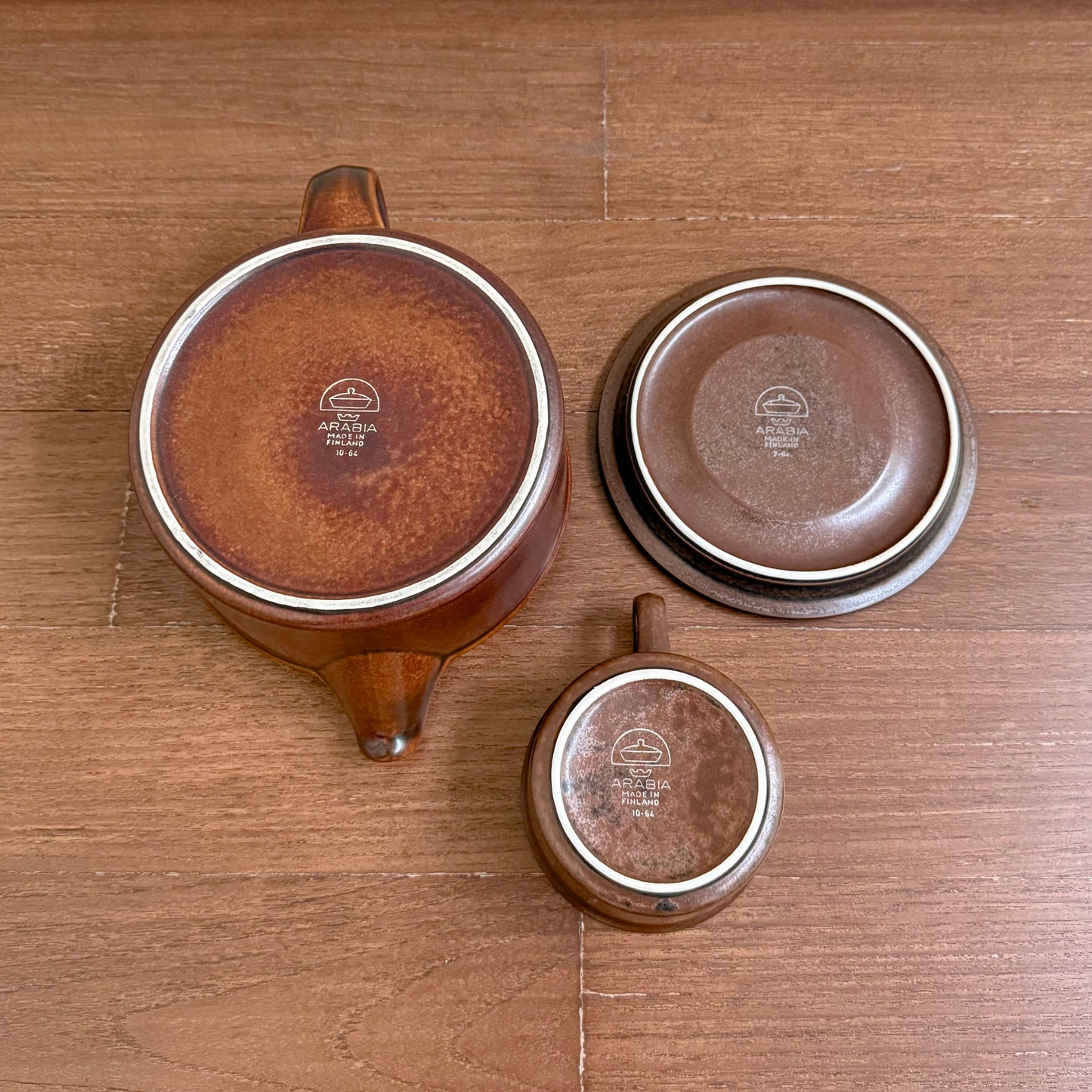 Bottom view of Arabia Finland Ruska Early Tea Set pieces arranged on a wooden surface, showcasing the vintage logo and warm brown ceramic glaze typical of Nordic drinkware.