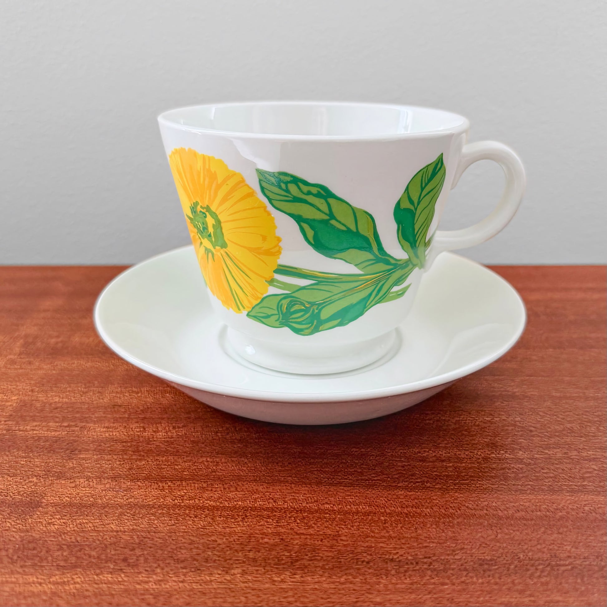 Arabia Finland Suvi Large Cocoa Cup with Saucer featuring a vibrant yellow flower and green leaves design on white ceramic, displayed on a wooden surface.