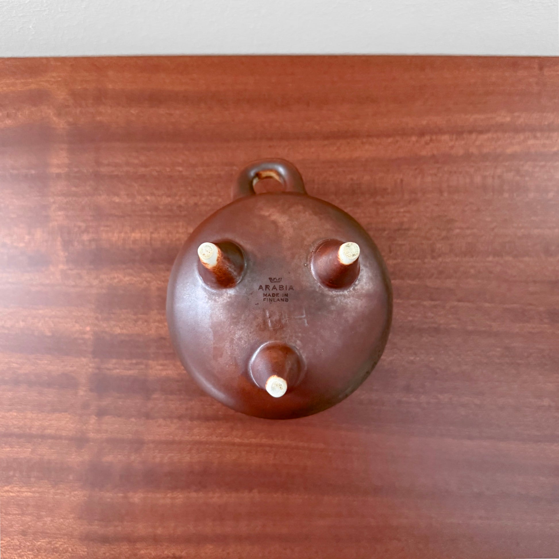 Brita Heilimo Arabia Finland Decorative Pot shown from the underside, revealing its three legs, handle, and signed vintage ceramic body on a wooden surface.